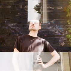 Doppelbelichtung: Portrait eines jungen Mannes, der eine Hand in die Hüfte gestemmt hat, vor einer dunkel bemalten Backsteinwand, zusammen mit Teilen zweier Frames mit brutalistischer Architektur, einer der Frames mit Passant