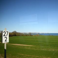 Blick über ein gr&uuml;nes Feld auf die leuchtend blaue Ostsee, links im Bild eine Kilometertafel