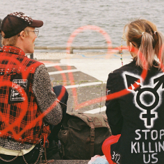 Farbfilmfoto zweier sitzender Punks, von hinten aufgenommen, auf der einen Jacke ein Trans-Symbol und die Worte "Stop killing us"