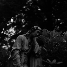 Recht dunkles Schwarzweiß-Film-Foto einer Statue, die ihren Kopf auf eine Hand stützt und den Ellbogen auf eine Säule. Um die Statue herum ist Rhododendron, im Hintergrund sind Bäume, mit viel Bokeh. 